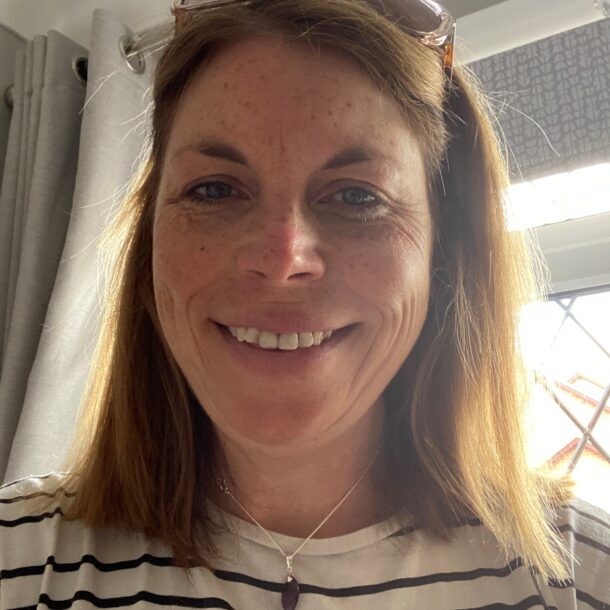 Heidi Abernethy A woman with shoulder-length light brown hair smiling indoors, wearing a white and black striped shirt, a purple pendant necklace, and sunglasses resting on her head.