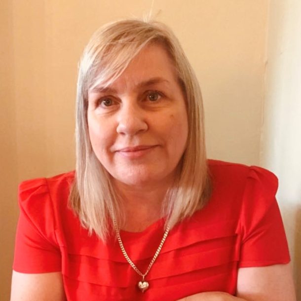 A person with light colored, shoulder-length hair wearing a bright red top with pleated detailing and a gold heart-shaped pendant necklace, seated indoors against a neutral background.