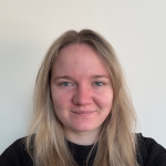 Close-up portrait of a person with long, light-coloured hair, looking at the camera against a plain light background, wearing a dark top and a small nose stud.