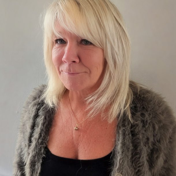 Close-up of a person with shoulder-length blonde hair wearing a dark top and grey jacket.